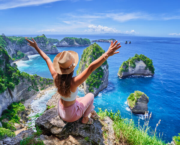 Woman traveler with open arms sitting on rock cliff and enjoying of scenic landscape of Diamond beach during vacation to Nusa Penida Island in Indonesia, travel to touristic beautiful destinations
